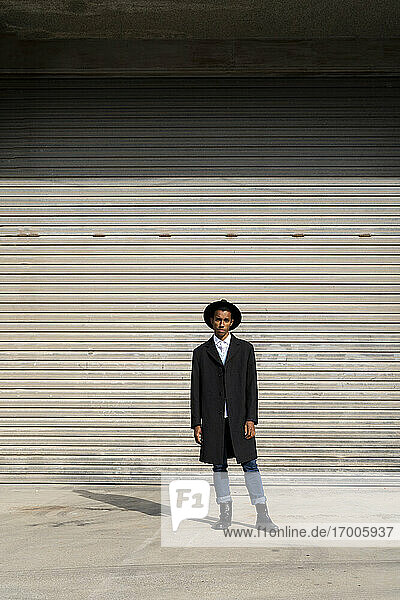 Modischer junger Mann  der an einem sonnigen Tag vor einem Fensterladen steht