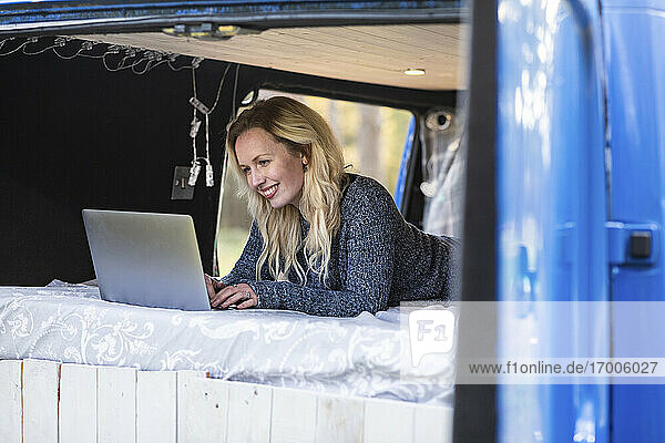 Lächelnde Frau  die einen Laptop benutzt  während sie auf einem Bett im Van liegt