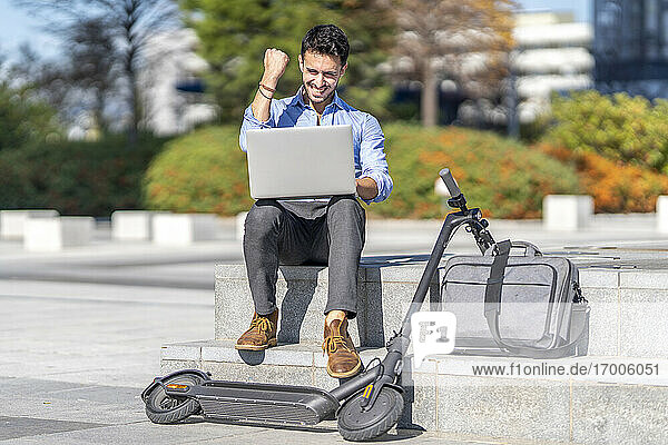 Smiling businessman showing winning gesture while sitting by briefcase and electric push scooter in city