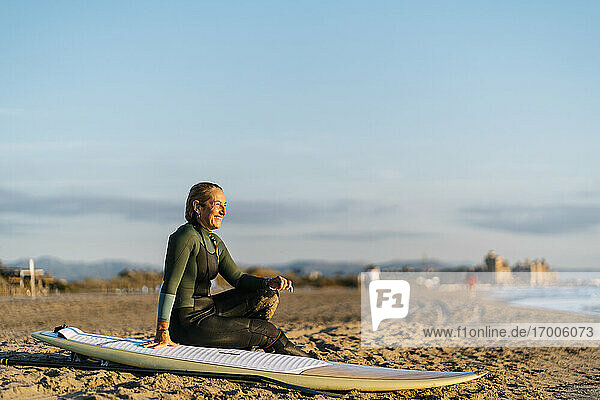 Smiling woman with paddleboard looking away while sitting on sand at beach during dawn