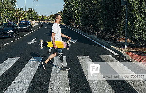 Junger Mann mit Beinprothese  der ein Skateboard hält und über einen Zebrastreifen geht