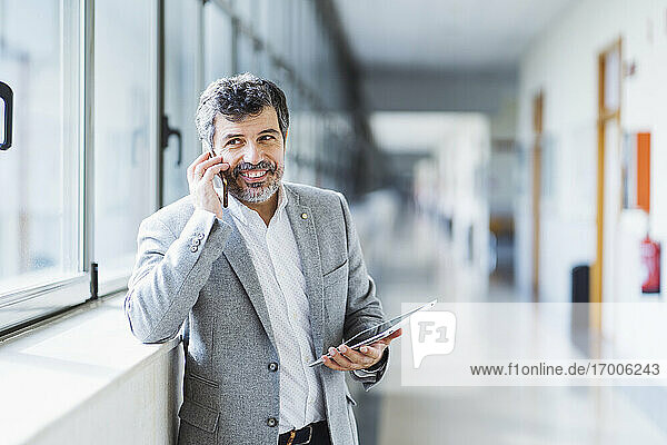 Lächelnder Professor  der wegschaut  während er im Flur der Universität das Telefon abnimmt