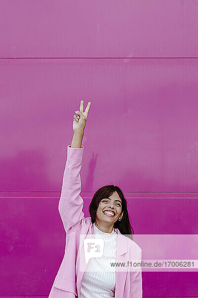 Lächelnde Frau mit erhobener Hand  die ein Friedenszeichen macht  während sie an einer rosa Wand steht