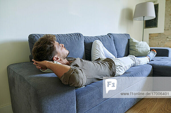Junger Mann entspannt sich auf dem Sofa mit den Händen hinter dem Kopf im Wohnzimmer