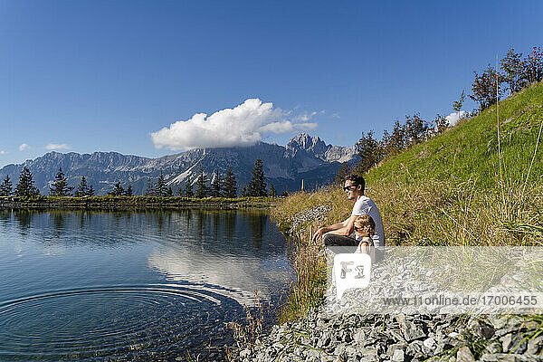 Vater sitzt mit kleiner Tochter am Ufer eines Alpensees