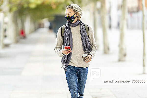 Junger Mann mit Gesichtsmaske  der beim Spaziergang in der Stadt ein Mobiltelefon benutzt