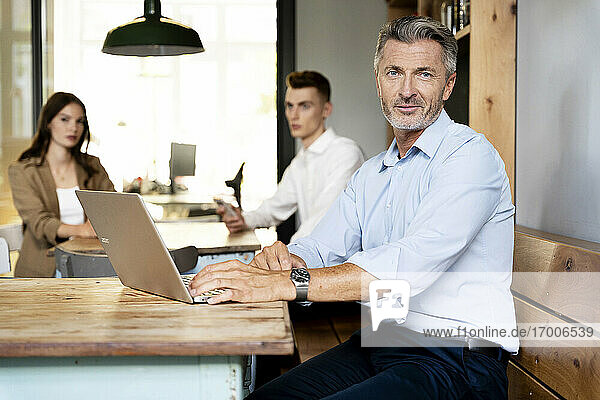 Älterer Geschäftsmann  der einen Laptop benutzt  während er mit Kollegen im Hintergrund im Büro sitzt