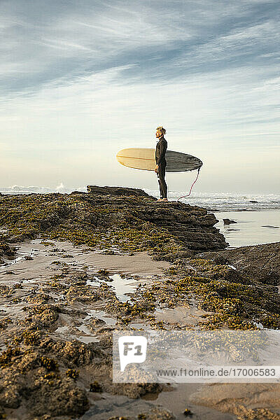 Male surfer standing on rock formation with surfboard at beach