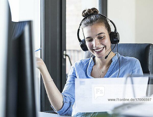 Geschäftsfrau mit Papier  die über Kopfhörer spricht  während sie im Büro sitzt