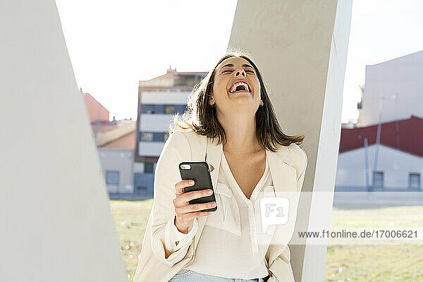 Weibliche Unternehmerin lacht  während sie ein Smartphone in der Stadt an einem sonnigen Tag hält