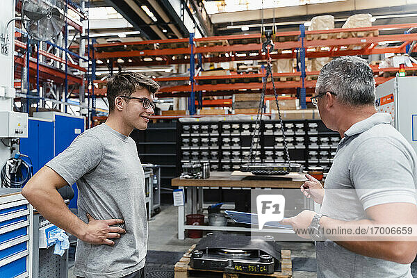 Lächelnder männlicher Mitarbeiter betrachtet einen reifen Ingenieur in einer beleuchteten Fabrik