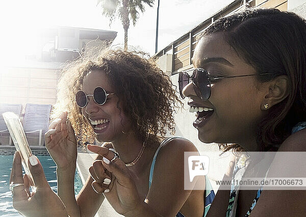 Cheerful young female friends sharing memories on smart phone at poolside