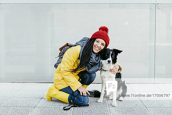 Smiling woman embracing dog while kneeling on footpath against wall