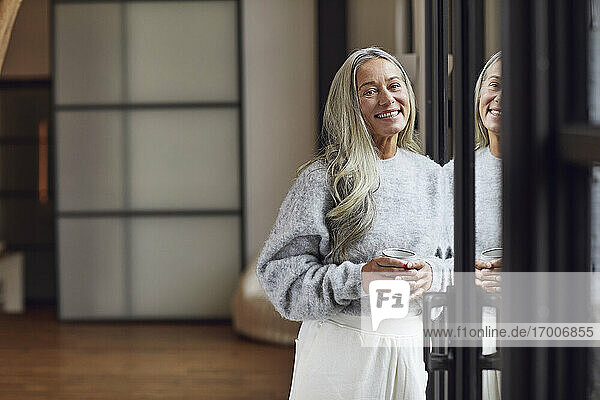 Lächelnde reife Frau mit grauen Haaren  die eine Kaffeetasse hält und sich zu Hause an die Tür lehnt
