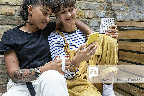 Lesbisches Paar mit Teetasse und Mobiltelefon im Hinterhof sitzend