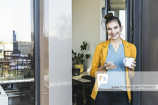Geschäftsfrau mit Kaffeetasse  die ein Mobiltelefon benutzt  während sie an der Tür im Büro steht