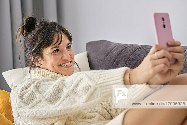 Lächelnde schöne Frau im Pullover nimmt Selfie durch Smartphone beim Entspannen zu Hause