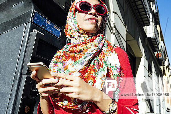 Frau in geblümtem Hijab schaut weg  während sie an einem sonnigen Tag ein Mobiltelefon hält