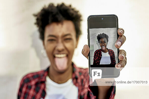 Fröhliche junge Frau mit herausgestreckter Zunge zeigt ihr Foto auf dem Smartphone
