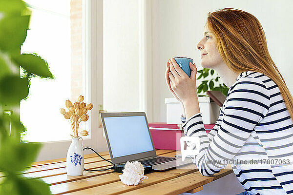 Junge rothaarige Frau mit Laptop trinkt Kaffee  während sie zu Hause sitzt