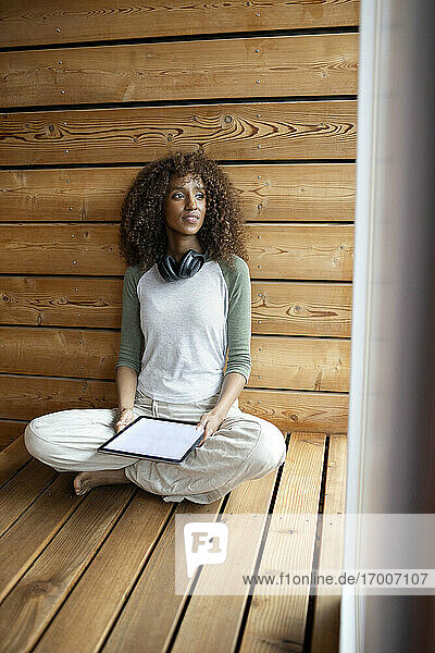 Junge Frau mit digitalem Tablet  die wegschaut  während sie im Schneidersitz zu Hause sitzt