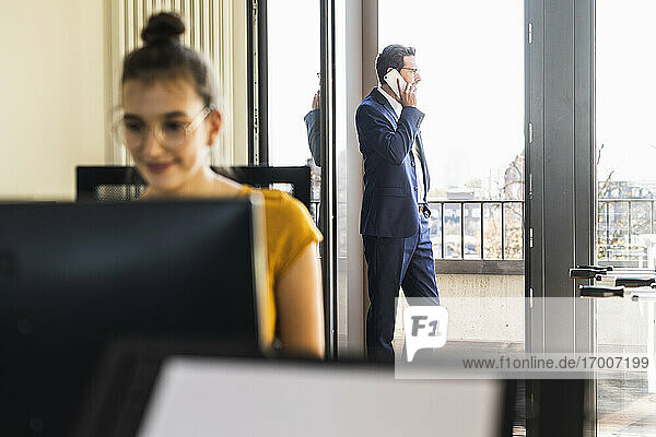 Ein Kollege arbeitet am Computer  während er im Büro sitzt und ein Geschäftsmann im Hintergrund auf dem Balkon steht