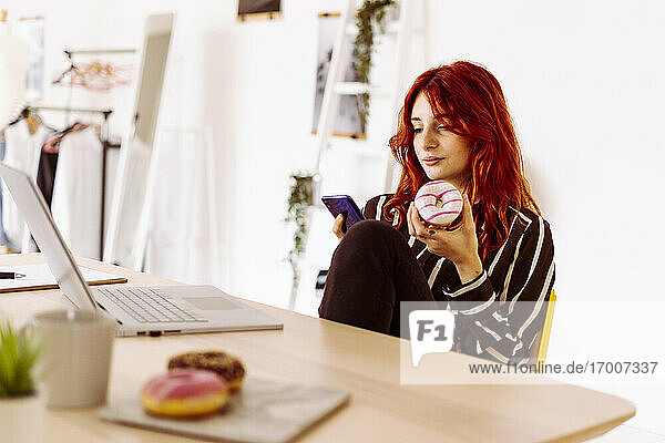 Junge Geschäftsfrau  die einen Donut in der Hand hält und ein Mobiltelefon benutzt  während sie im Büro sitzt