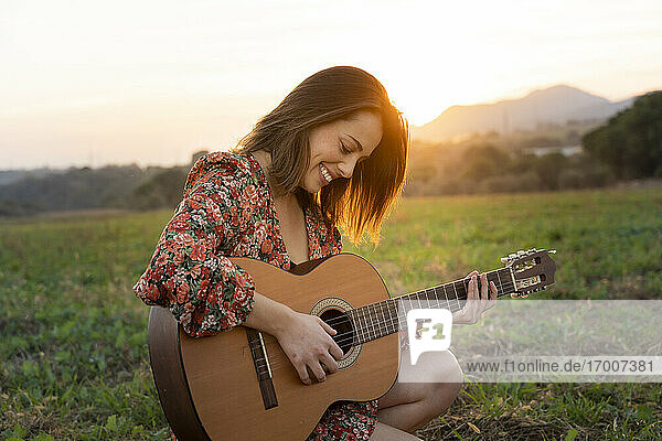 Lächelnde junge Frau spielt Gitarre auf einer Wiese bei Sonnenuntergang