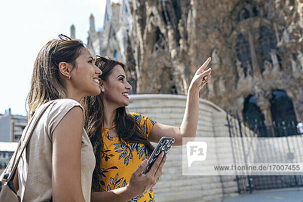 Smiling woman pointing while standing by friend using mobile phone at Sagrada Familia at Barcelona  Catalonia  Spain