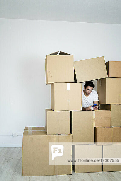 Junger Mann benutzt sein Smartphone  während er sich auf Kisten in einem neuen Haus stützt