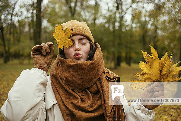 Young woman with eyes closed puckering while covering eye with autumn leaf in public park