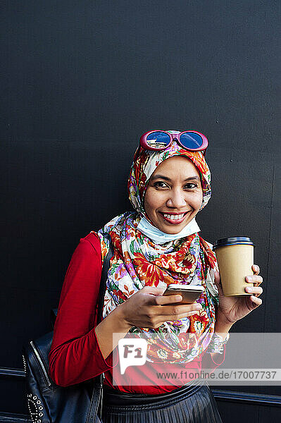 Lächelnde Frau mit Hidschab  die ein Handy und eine Kaffeetasse gegen eine schwarze Wand hält  während einer Pandemie