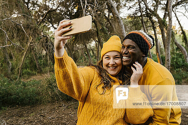 Lächelnde Freundin  die ein Selfie mit ihrem Freund durch ein Handy macht  während sie im Wald steht