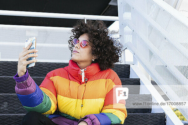 Woman wearing colorful jacket taking selfie through mobile phone while sitting on steps