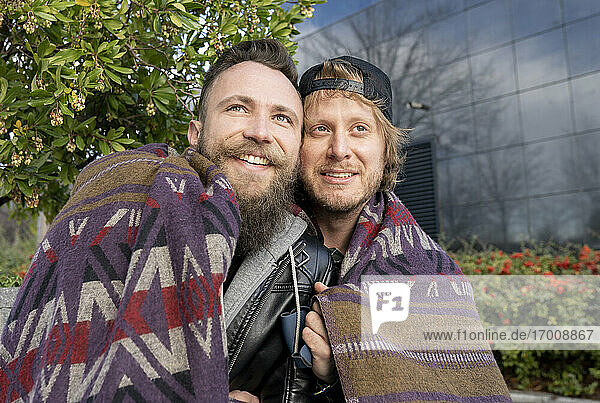 Close-up of smiling gay couple wrapped in shawl while sitting outdoors