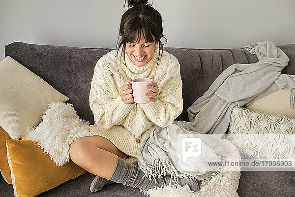 Glückliche Frau mit Kaffee im Becher  während sie im Winter zu Hause auf dem Sofa sitzt