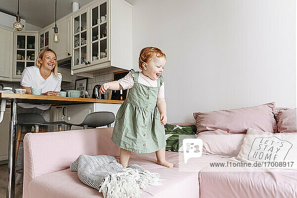 Kleines Mädchen springt auf dem Sofa  lächelnde Mutter im Hintergrund
