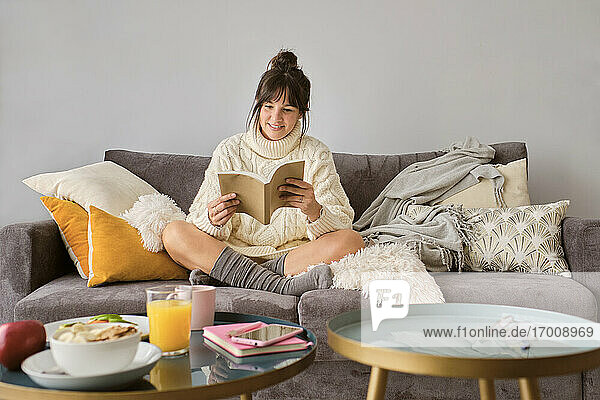 Lächelnde Frau im Pullover liest ein Buch  während sie zu Hause auf dem Sofa sitzt