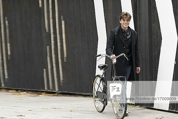 Smiling young man wheeling bicycle on footpath by wall