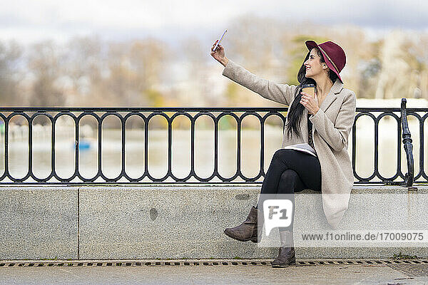 Mittlere erwachsene Frau  die einen Kaffee hält und ein Selfie mit ihrem Smartphone macht  während sie auf einer Stützmauer sitzt