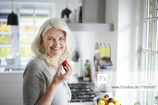 Lächelnde ältere Frau mit langen weißen Haaren  die eine frische Erdbeere in der Hand hält  während sie in der Küche zu Hause steht
