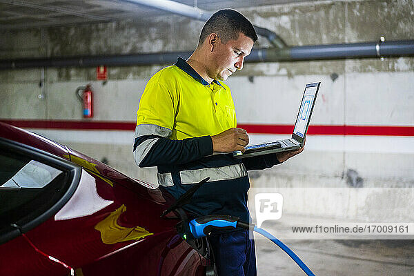 Männlicher Techniker  der ein Mobiltelefon mit Laptop an einem Elektroauto in einer Autowerkstatt benutzt