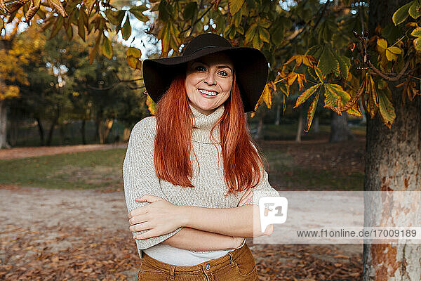 Lächelnde schöne Frau mit Hut und verschränkten Armen im Herbst im Park stehend
