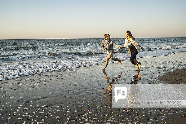 Glückliches Paar hält Hände beim Laufen am Strand gegen den klaren Himmel bei Sonnenuntergang