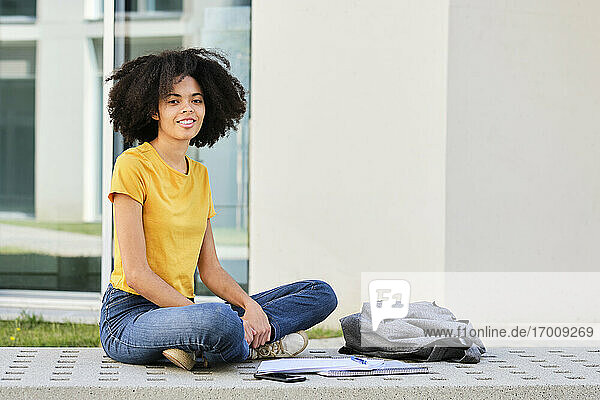 Junge Studentin sitzt im Schneidersitz bei Büchern und Tasche auf dem Campus
