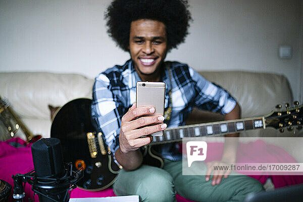 Lächelnder junger männlicher Gitarrist  der zu Hause mit seinem Smartphone Videogespräche führt