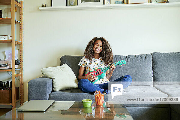 Smiling young woman with ukulele sitting on sofa in living room