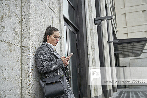 Businesswoman using smart phone while leaning on building wall