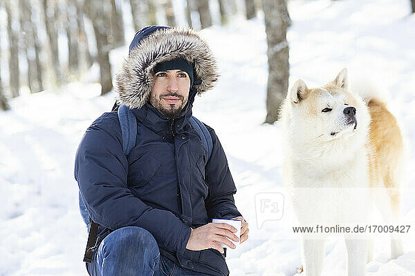 Mid adult man and akita dog looking away while holding coffee cup in snow