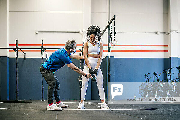Male fitness in mask instructor explaining sportswoman about kettlebell in gym
